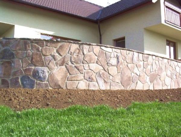pohled na rodinný dům, na jehož pohled překrývá divákovi kamenná zeď tvořená nepravidelným kamením nepravidelných tvarů a velikostí.